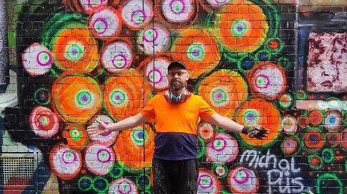 "Incubator Burst" - Neon Fluorescent Roundism Mural by Michal Plis (2026) Street art mural "Incubated Burst" by Michal Plis, painted on an alley brick wall. The piece features an organic, energetic cluster of glowing concentric circles in vibrant neon orange, lime green, and magenta, symbolizing cellular incubation, spring, and